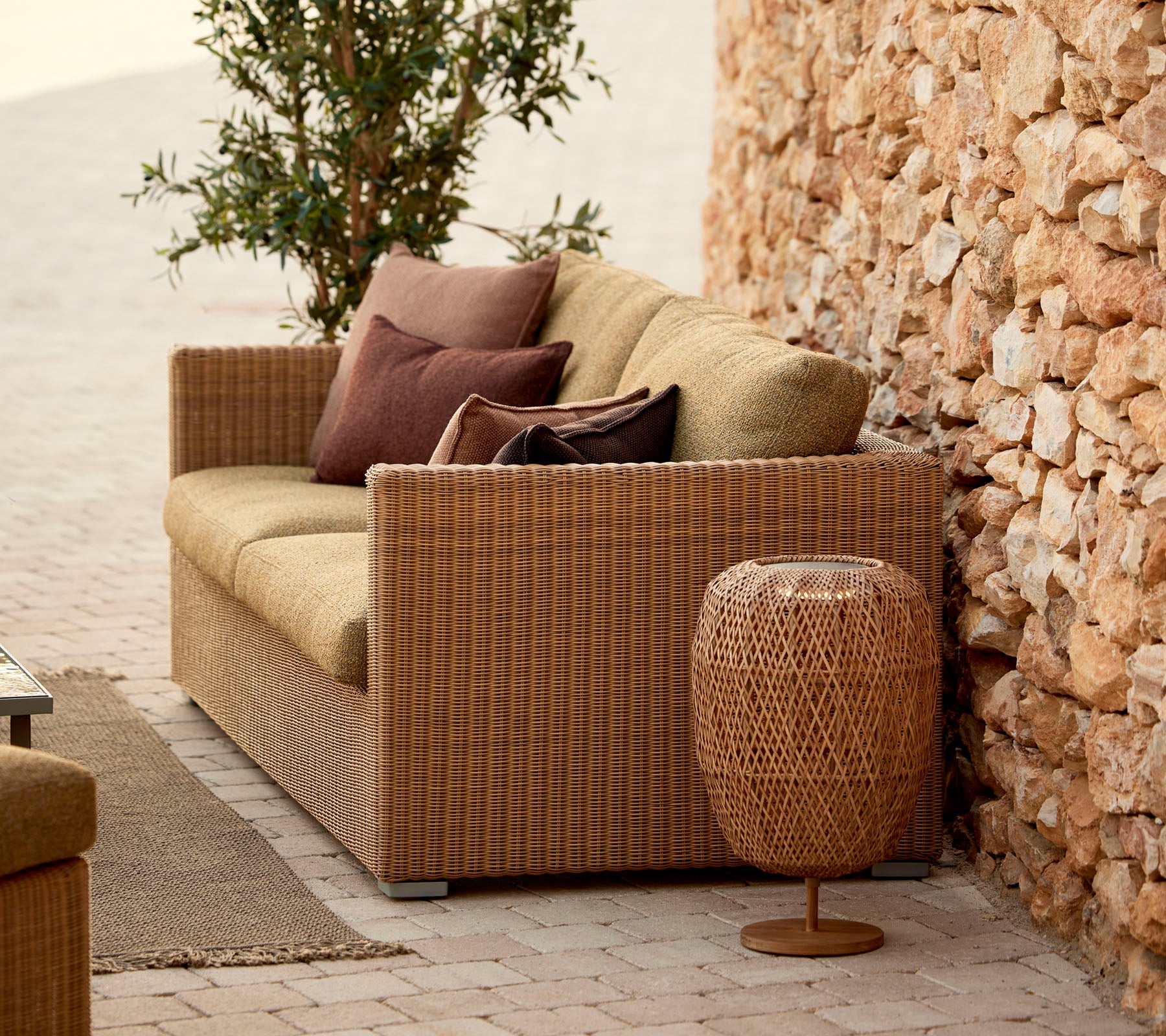 A cozy wicker sofa with pillows, complemented by a stylish woven side table, set against a stone wall and lush greenery.
