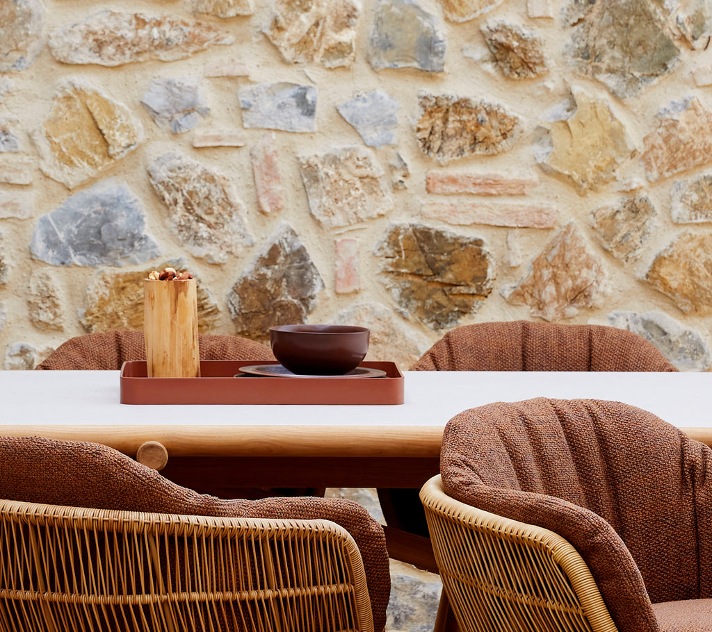 A cozy arrangement featuring a desert red club tray, a brown bowl, and stylish woven chairs against a stone wall backdrop.