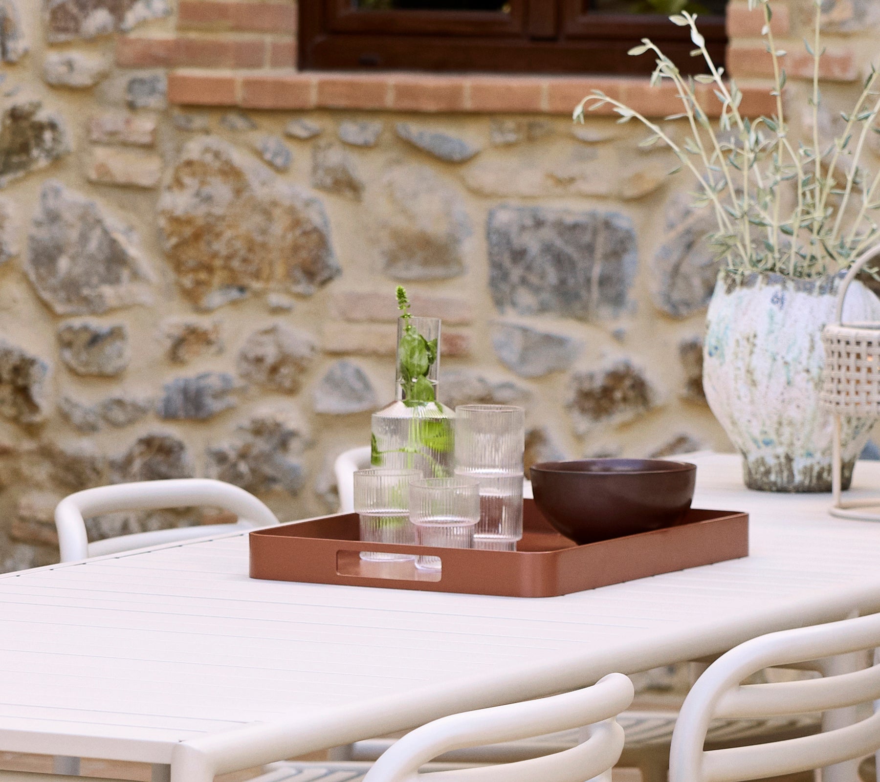 A club tray in desert red holds glasses and a bowl, accompanied by a decorative vase and greenery against a stone backdrop.
