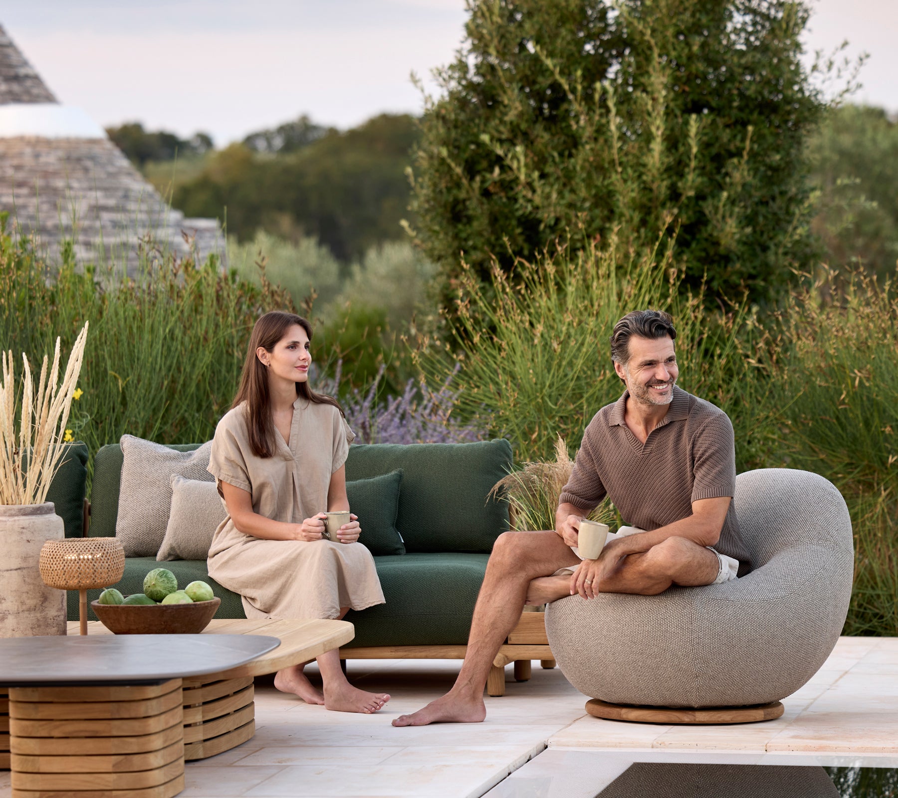 Modern lounge with green sofa, grey rounded chair, and brown table surrounded by greenery.