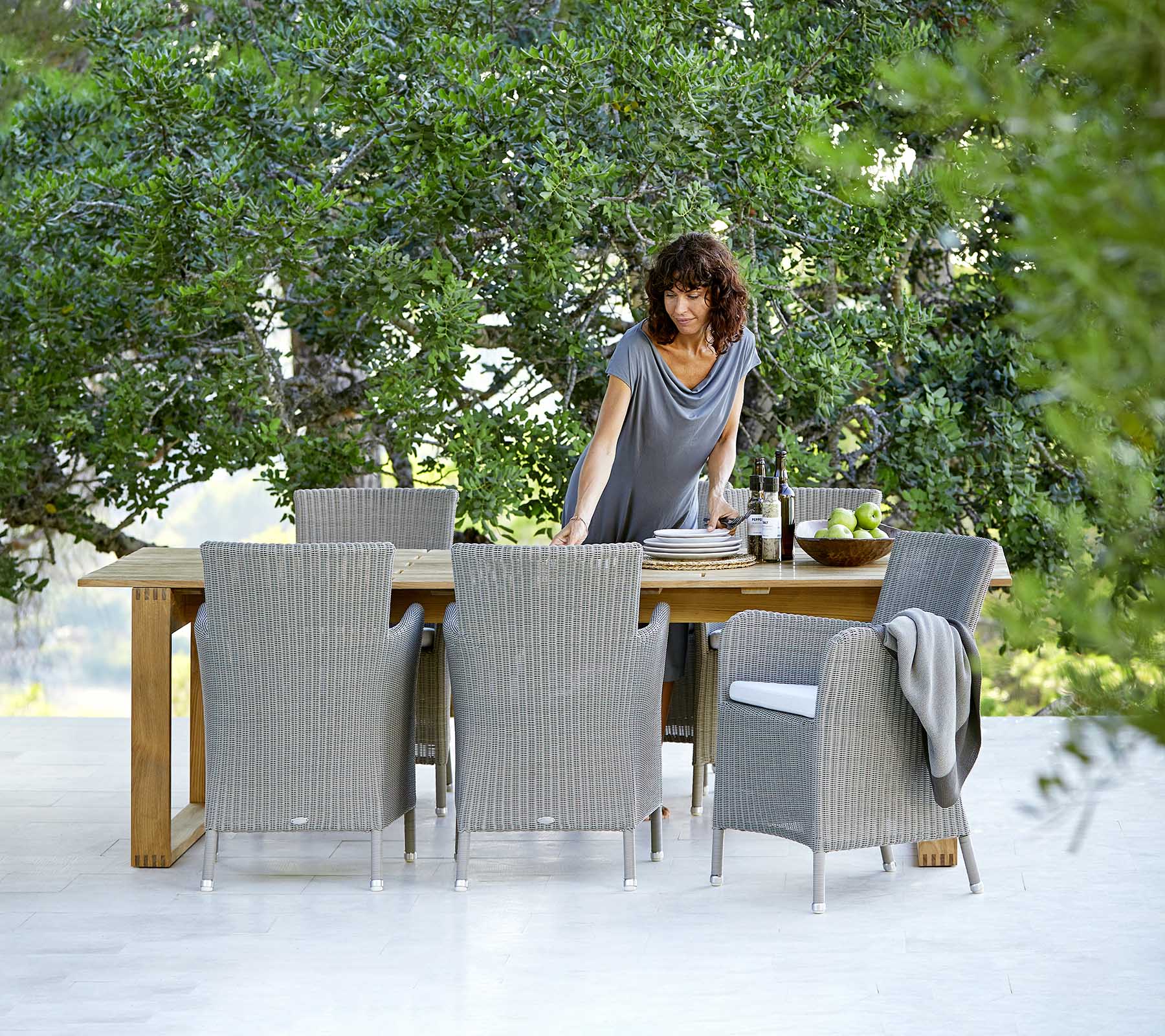 A woman interacts with a dining table surrounded by wicker chairs, set against a backdrop of lush greenery.