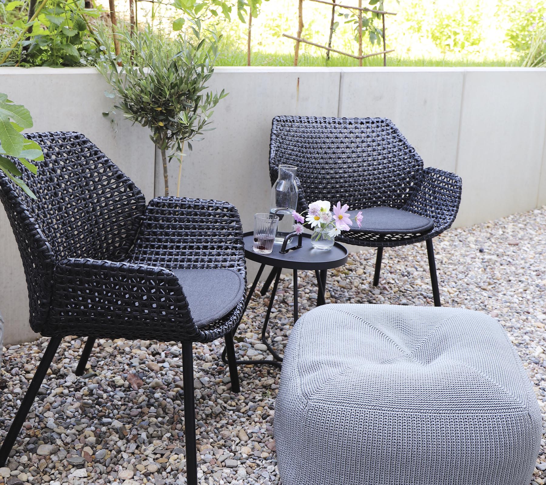 Two stylish black chairs paired with a small table, adorned with a vase of flowers and a cozy ottoman on a gravel surface.