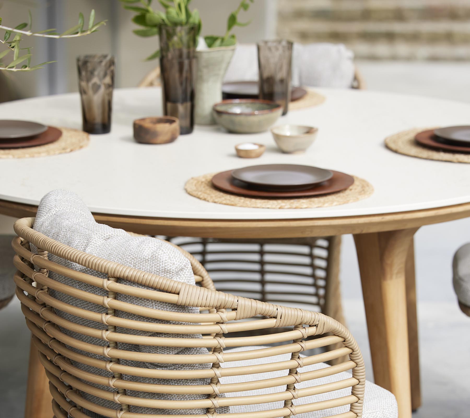 A stylish dining table set with plates, bowls, and decorative glasses, surrounded by woven chairs and natural elements.