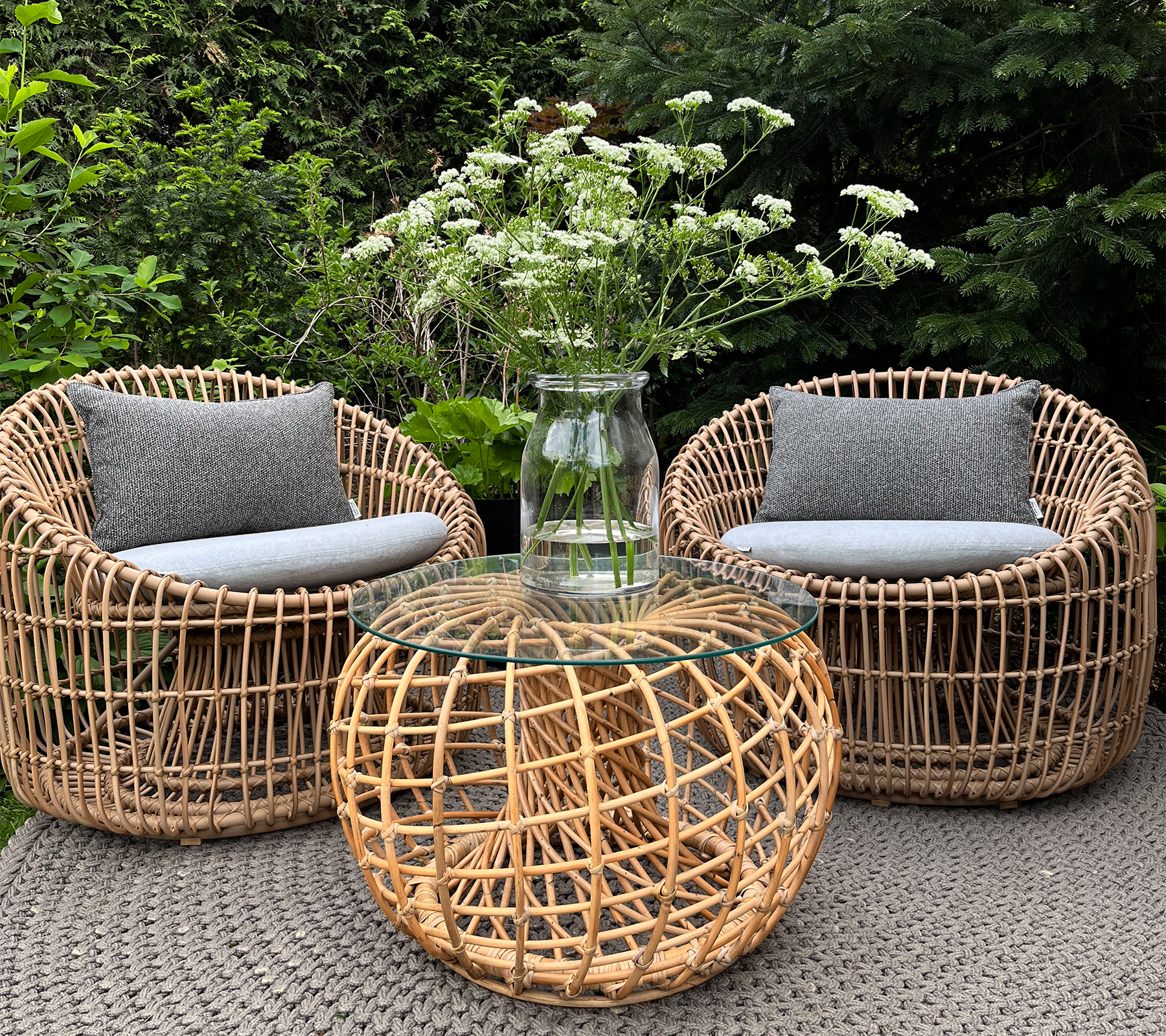 Two woven chairs with gray cushions surround a round woven coffee table topped with a clear vase holding white flowers.