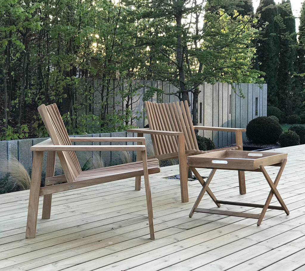 Two wooden chairs and a small folding table, set against a backdrop of greenery and wooden panels. Natural and inviting outdoor seating.