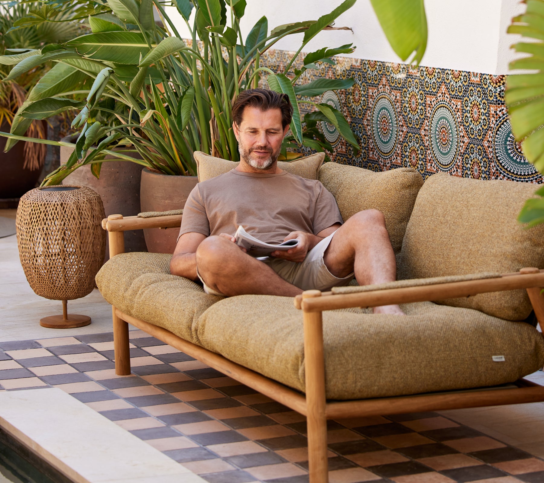 A man relaxes on a comfortable couch, reading a magazine, surrounded by lush greenery and decorative patterns.