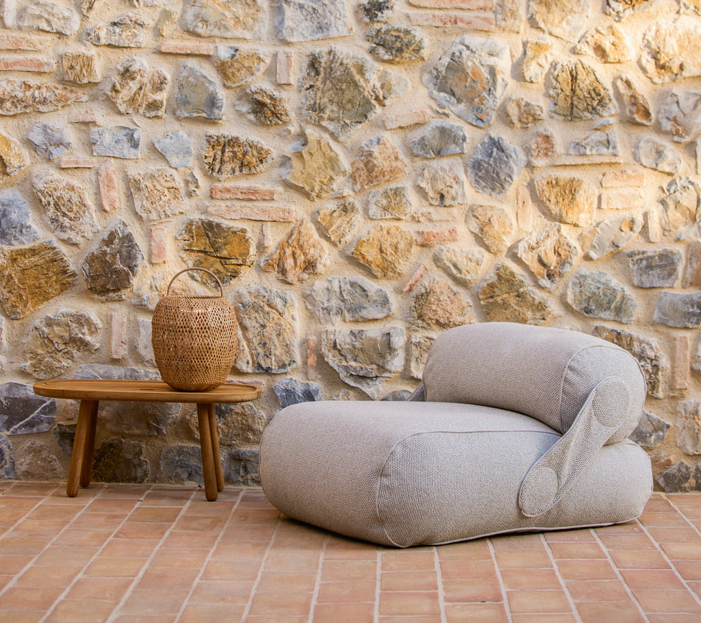 A cozy lounge chair beside a woven basket on a wooden table, with a textured stone wall in the background.