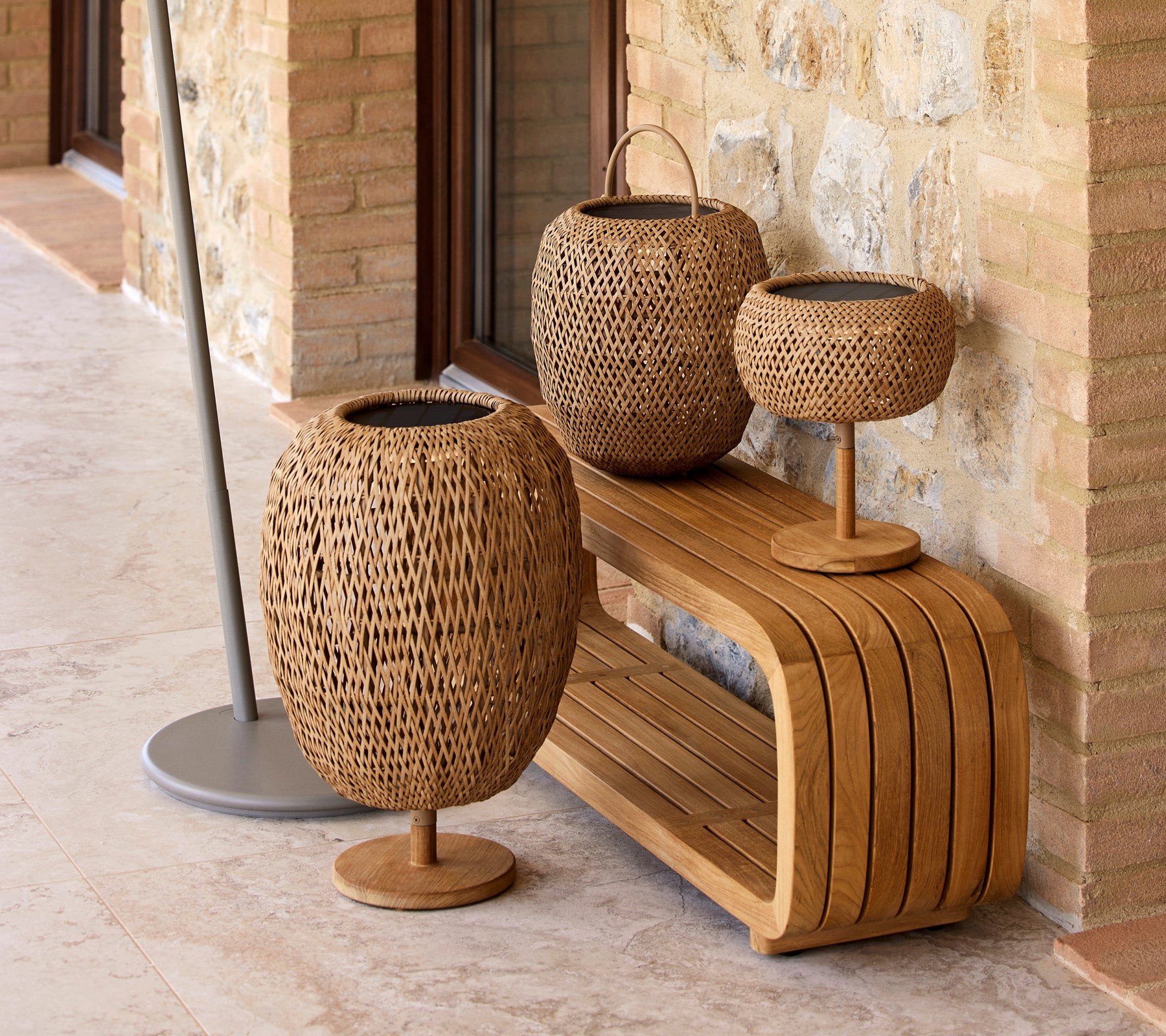 Three woven lamps from the Illusion Glow series arranged on a stylish wooden bench, set against a stone wall.