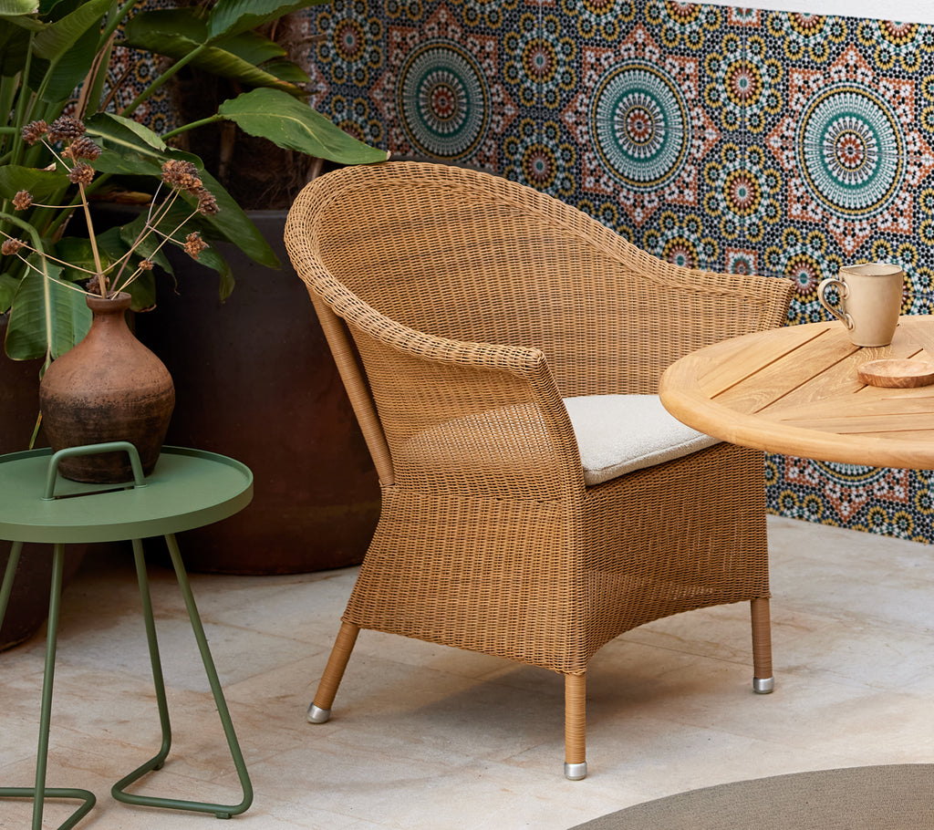 A wicker chair with a cushion beside a green side table, featuring a decorative pattern in the background and a coffee cup on the table.
