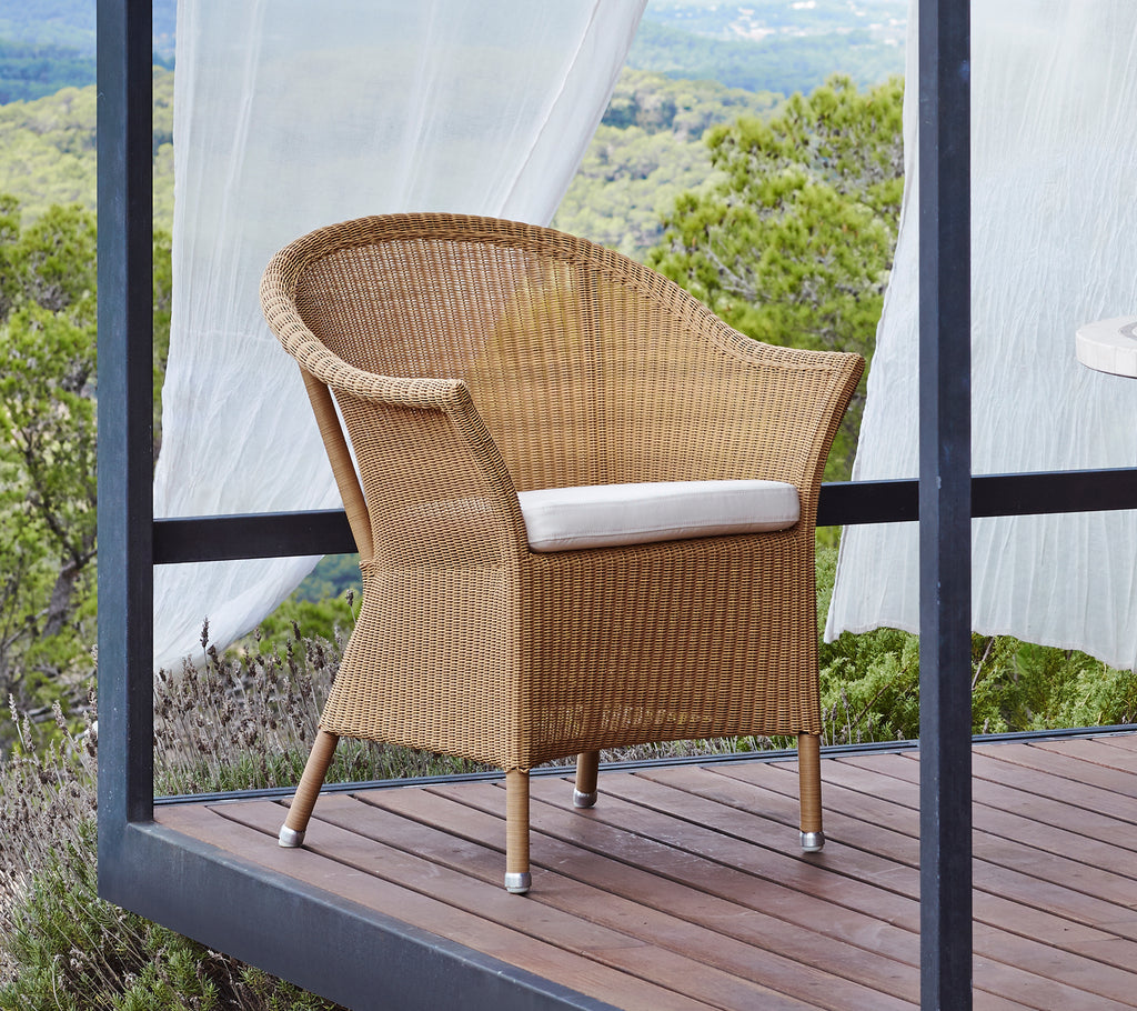 Natural woven chair with a comfortable white cushion, featuring curved arms and sturdy legs, set against a scenic backdrop.