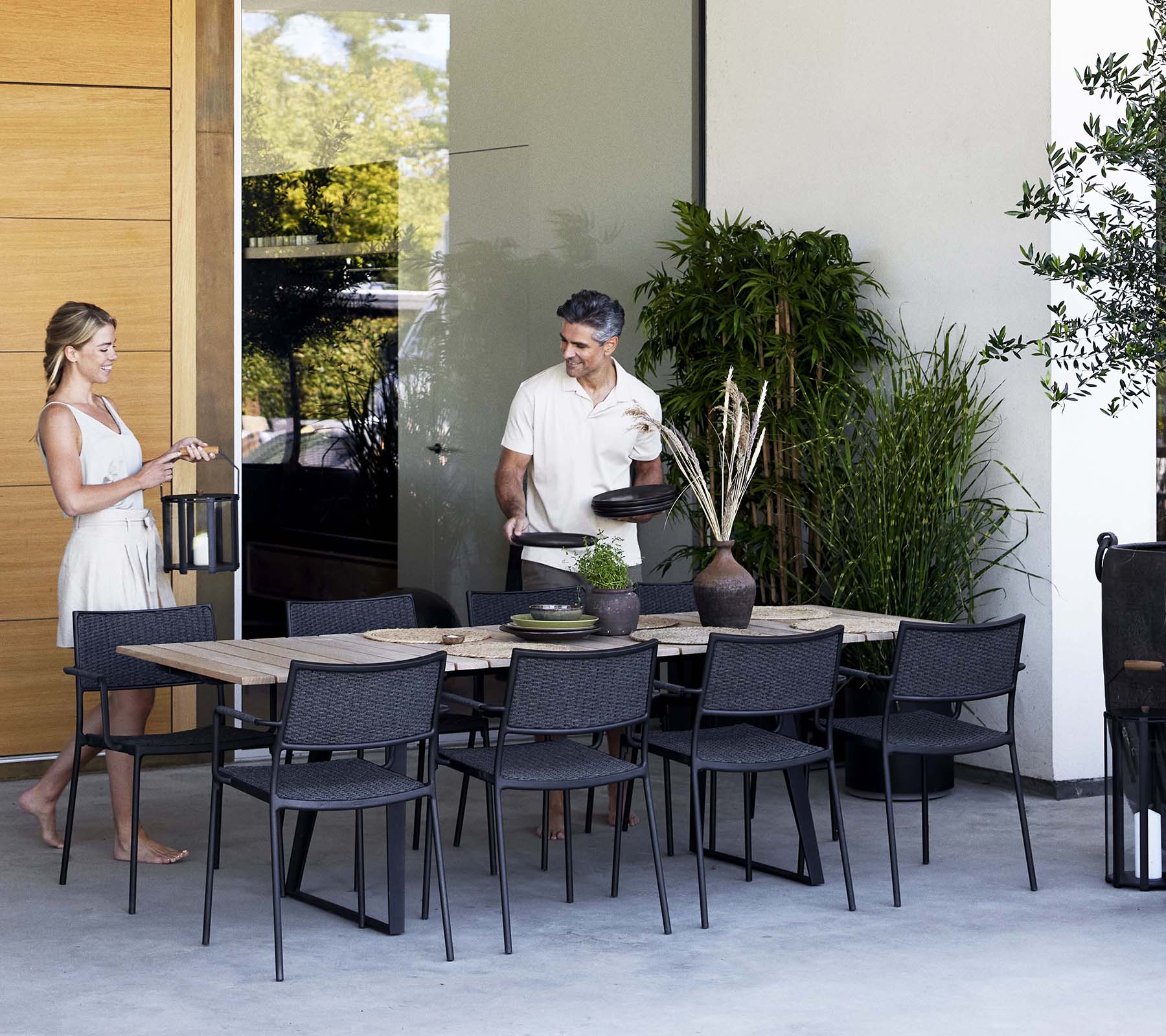 A woman and a man engage in conversation near a stylish dining table surrounded by modern chairs and greenery.