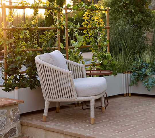 Moments lounge chair in sand color beside a planter with greenery, creating a cozy and inviting seating area.