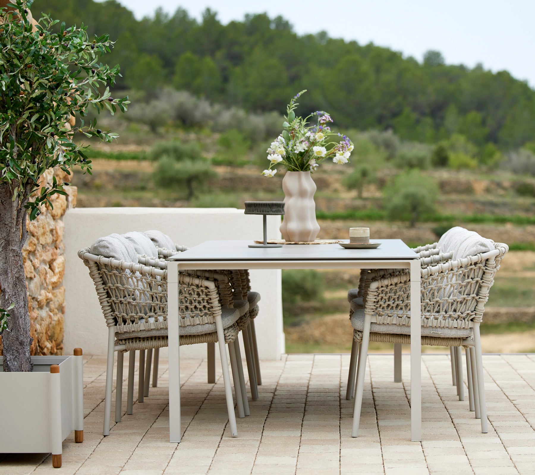 A stylish outdoor dining table set with four woven chairs, adorned with a vase of flowers and tabletop accessories, surrounded by greenery.