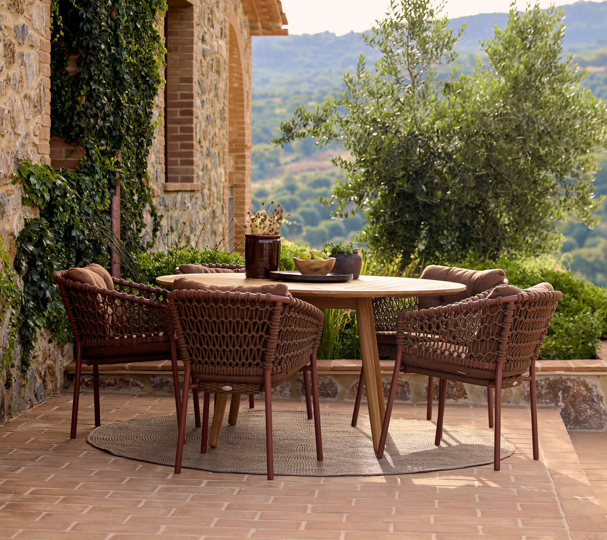 A round dining table surrounded by four umber brown Ocean dining chairs, set against a lush outdoor backdrop.