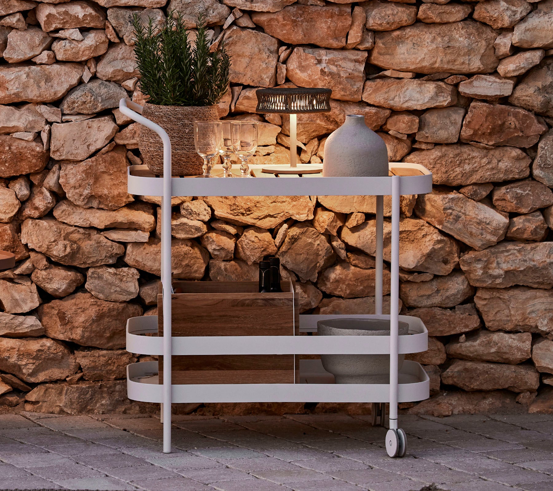 A stylish cart featuring a wooden top, white frame, decorative pot, and sleek lighting against a stone wall backdrop.