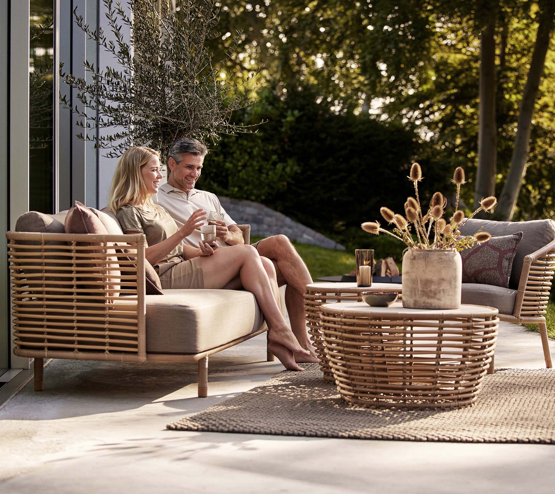 A couple enjoys drinks on a Sense sofa and lounge chair, accompanied by a stylish basket coffee table and decorative vase.