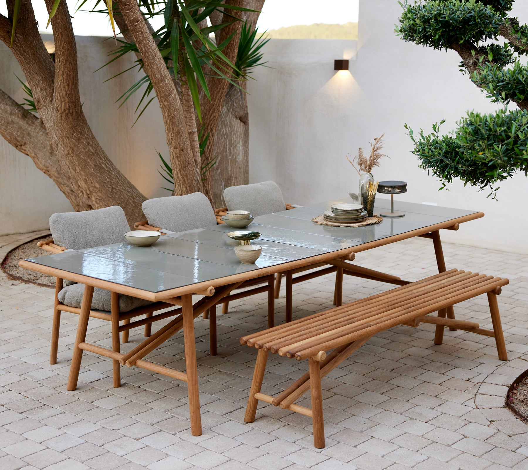 Teak dining table with glass top, accompanied by a bench and four upholstered chairs, set with dishes and ambient lighting.