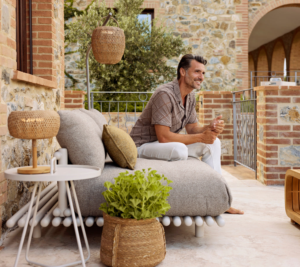 A man relaxes on a cozy, textured lounge with decorative cushions, beside a sleek side table and a warm, glowing lamp.