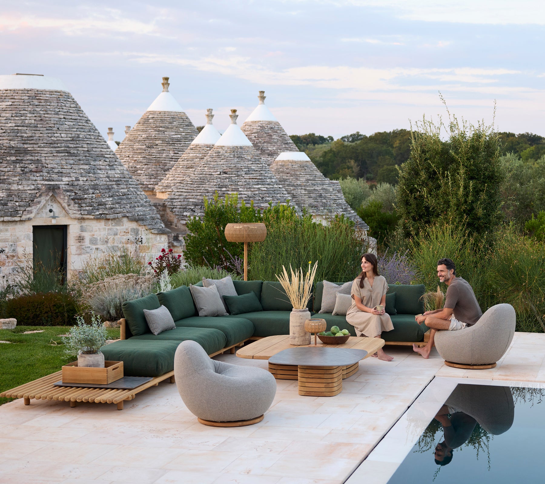 Modern outdoor lounge area with green sectional sofa and grey chairs by a pool.