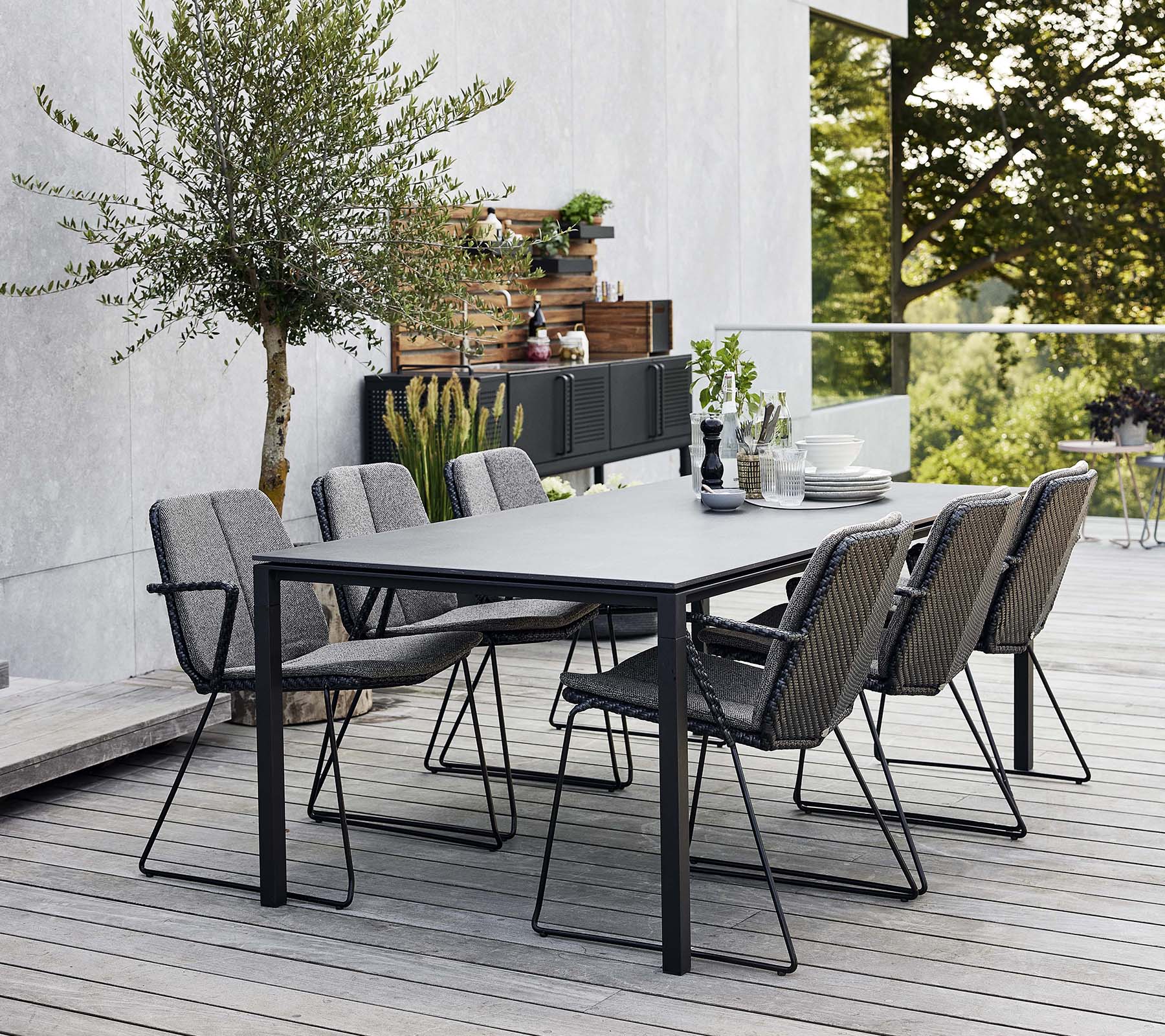 A stylish outdoor dining set featuring a sleek table and several comfortable armchairs, surrounded by greenery and a modern background.