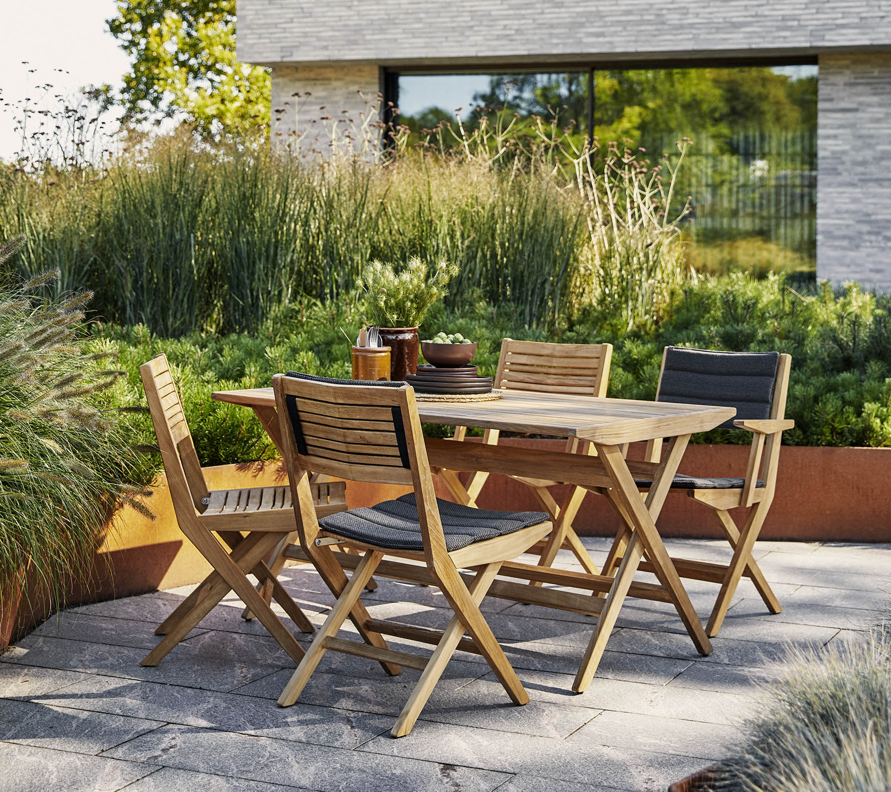 Stylish wooden table with matching chairs surrounded by lush greenery and a modern architectural backdrop. Perfect for outdoor gatherings.