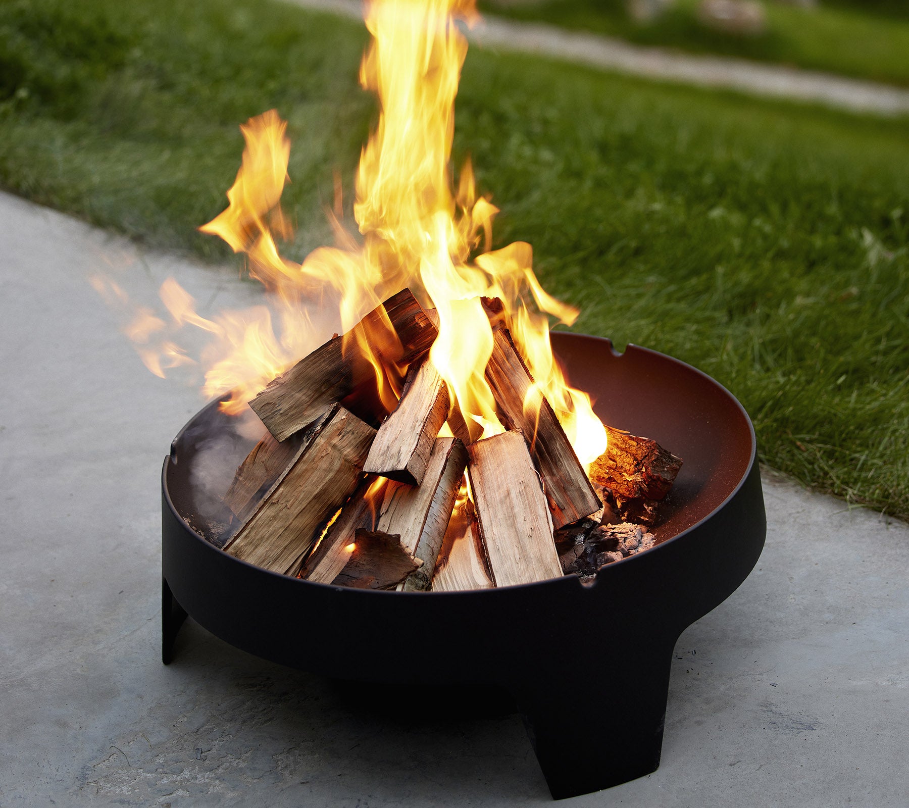 A fire pit with flames rising over stacked logs, surrounded by grass, creating a warm and inviting atmosphere.