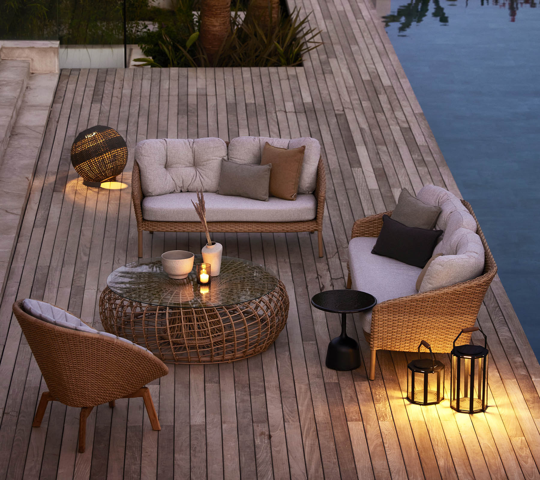 Cozy outdoor seating area with woven furniture, soft cushions, and ambient lighting beside a pool on a wooden deck.
