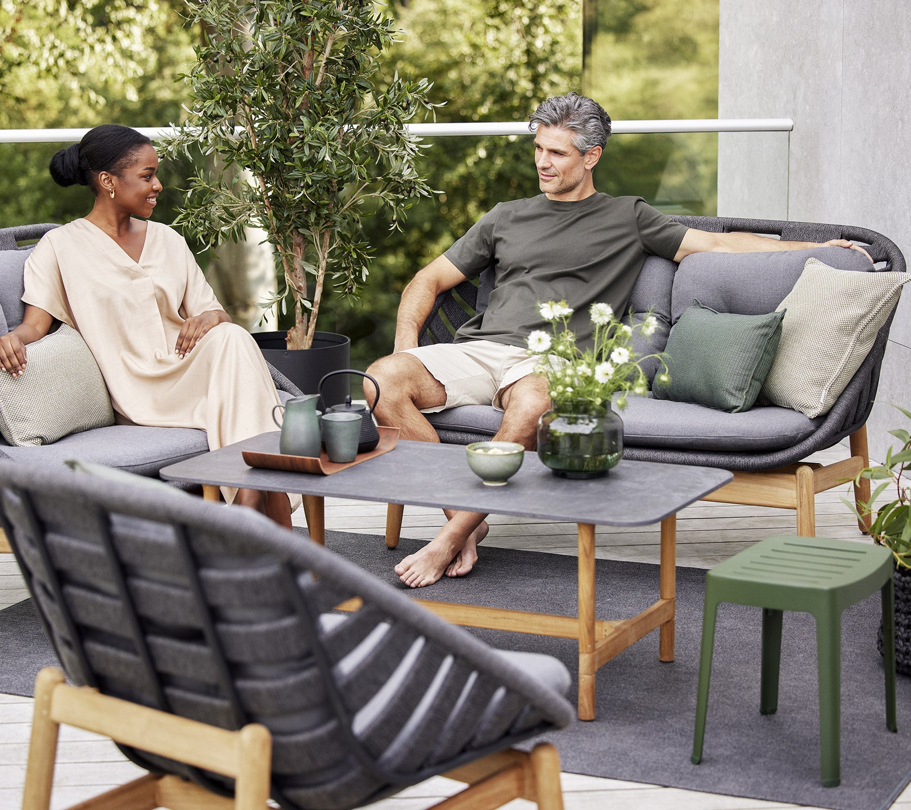 Two people relax on a stylish outdoor sofa, surrounded by greenery and decorative plants, enjoying a peaceful moment together.